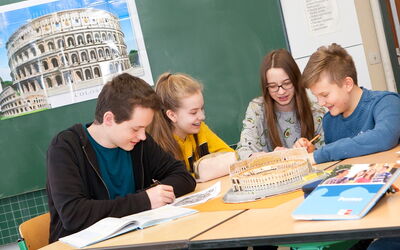 Vier SchülerInnen sitzen an einem Tisch vor einer Tafel und arbeiten gemeinsam an ihrem Projekt.