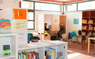 Ein weißes Bücherregal in der Schulbibliothek mit einer Sitzecke eigens für die Hausaufgabenbetreuung, in der zwei Schüler sitzen.