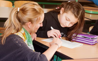 Eine Lehrerin sitzt neben einem Tisch und hilft einem Mädchen bei ihrer Aufgabe.