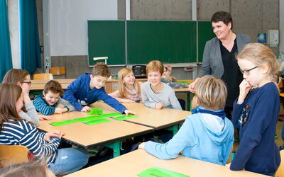 Eine Lehrerin unterrichtet eine Gruppe von Kindern in einem Klassenzimmer, alle hören aufmerksam zu.